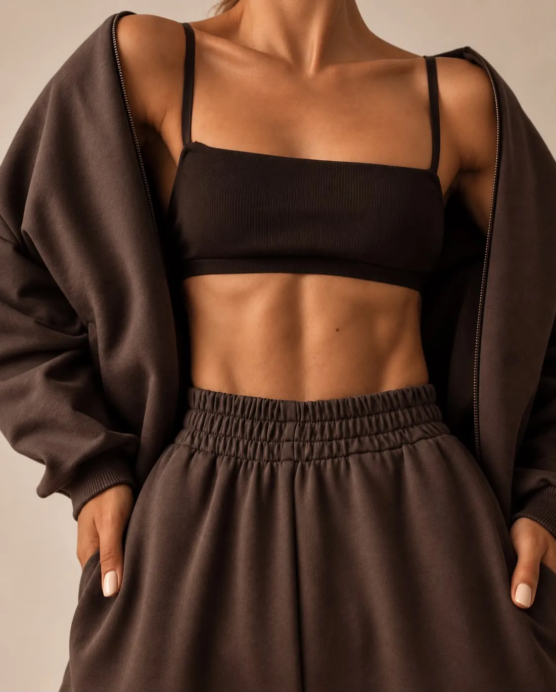 Buste en brassière noire et gilet de molleton brun ouvert, mains dans les poches, sangle abdominale dessinée, sur fond beige.