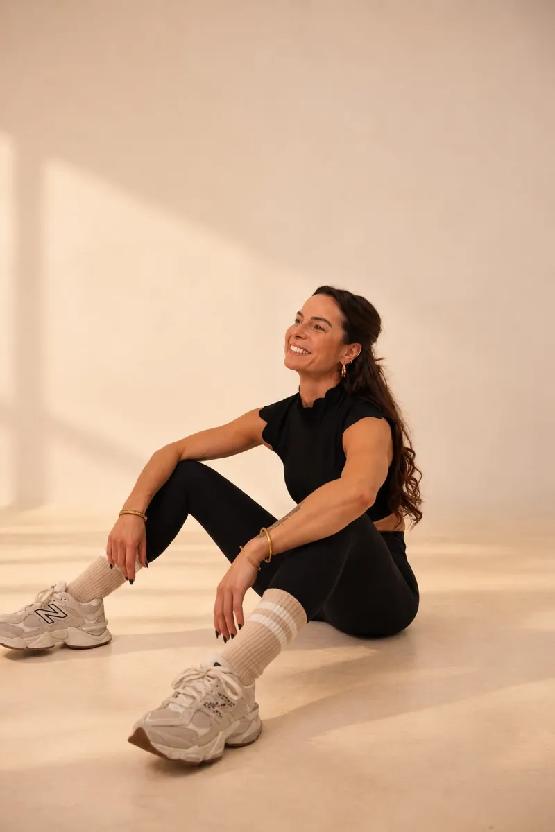 Lucile assise au sol, souriante, en tenue de sport noire, dans un studio baigné de lumière chaude