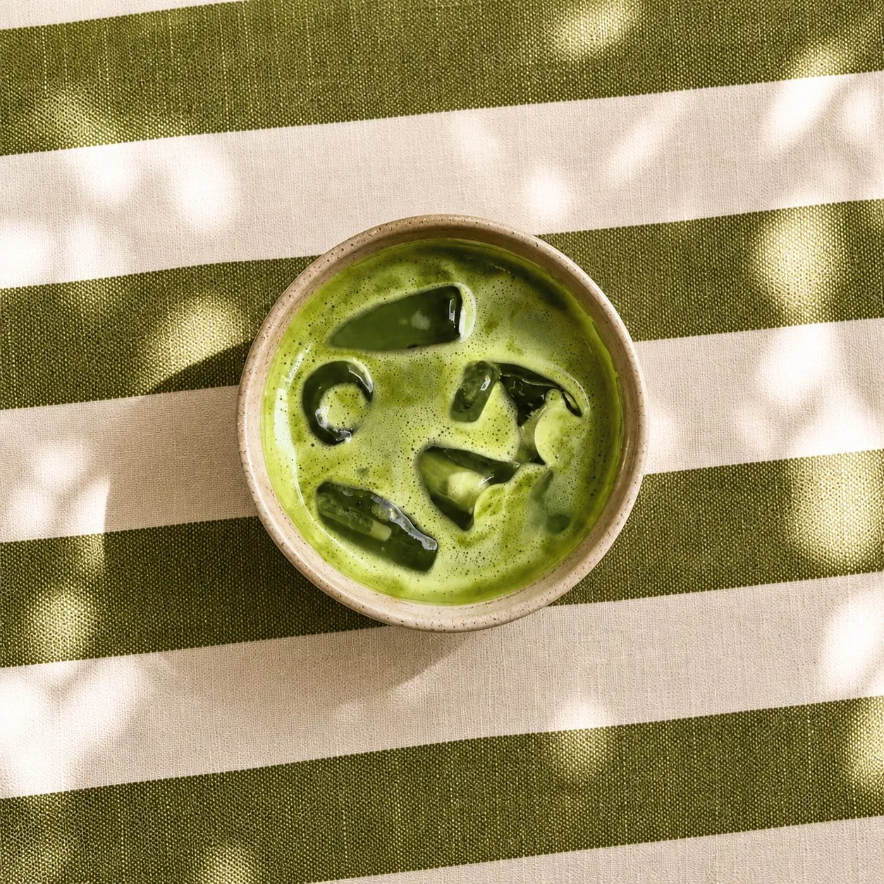 Bol en céramique de boisson glacée au matcha posé sur une nappe rayée vert olive et blanc, ombres de feuillage sur la table.