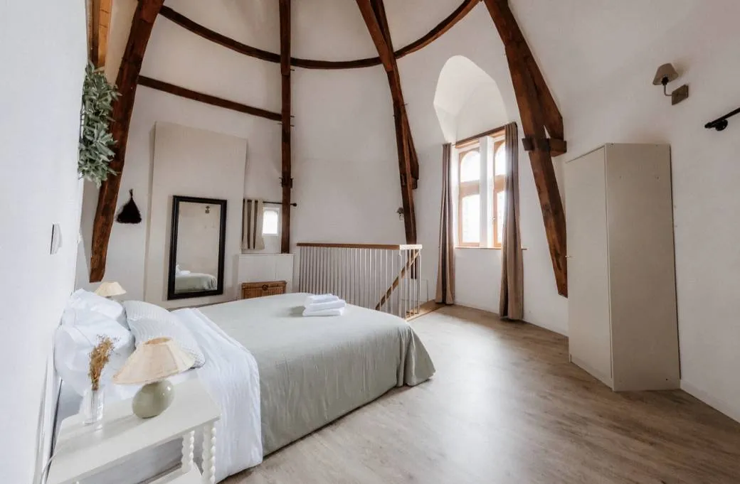 Une chambre du pavillon sous charpente apparente : lit double en linge blanc, murs clairs, parquet et fenêtre en arc.